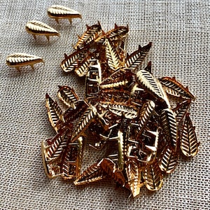 以下が含まれることがあります： 金色の金属製の葉の形をした装飾品がたくさん積まれています。装飾品はランダムなパターンで配置されており、表面はテクスチャ加工されています。