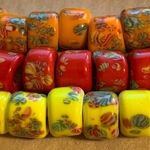 May include: Close-up of colorful glass beads in shades of orange, red, and yellow. The beads have a rectangular shape with rounded edges and decorative patterns in various colors. The beads are arranged in rows.