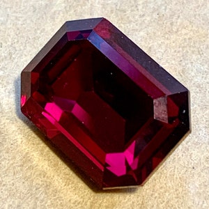 Puede incluir: Una piedra preciosa facetada de color rojo intenso con talla esmeralda. La piedra preciosa está engastada en un engaste de plata.