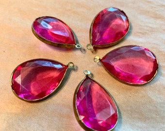 Puede incluir: Cinco colgantes de cristal rosa en forma de lágrima con engastes de metal dorado. Los colgantes tienen un diseño facetado y están dispuestos en círculo.