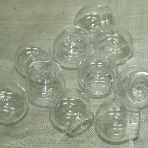 May include: A group of ten clear glass round jars with a rounded bottom. The jars are arranged in a pile on a light brown textured surface.