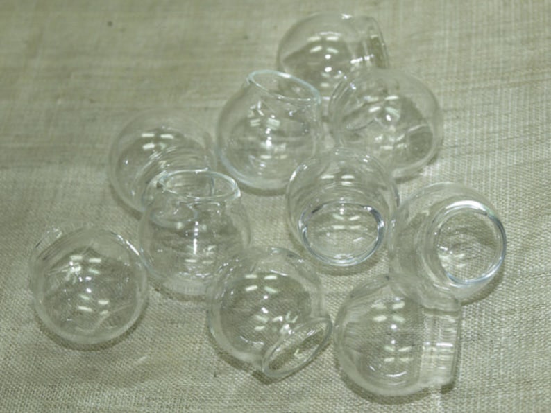 May include: A group of ten clear glass round jars with a rounded bottom. The jars are arranged in a pile on a light brown textured surface.