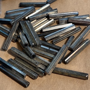 May include: A collection of silver-colored, hexagonal metal dowels scattered on a brown surface. The dowels are of varying lengths and appear to be made of a metallic material. The lighting creates highlights and shadows on the surfaces.