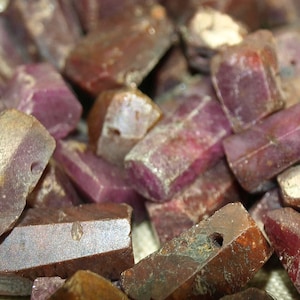 May include: A close-up of a pile of rough, unpolished, reddish-purple gemstones. The stones are rectangular in shape and have a natural, raw appearance.