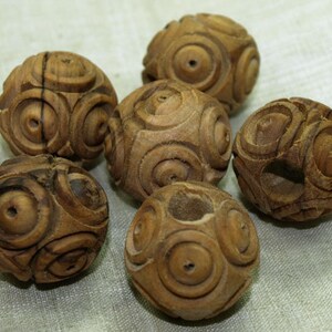 May include: Six small, intricately carved wooden spheres. Each sphere features a repeating pattern of concentric circles and a natural wood grain finish. The spheres are arranged on a light-colored surface, showcasing the detailed craftsmanship.