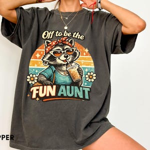 Puede incluir: Una camiseta gris carbón de manga corta con un gráfico de un mapache con gafas de sol y un pañuelo, sosteniendo un café helado. El texto dice Off to be the FUN AUNT. El diseño incluye un fondo de atardecer retro y flores blancas.