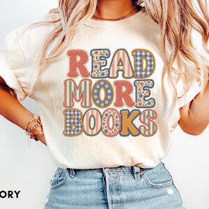 Puede incluir: Una camiseta de color marfil con el texto READ MORE BOOKS en letras grandes y gruesas. Cada letra está rellena con un patrón diferente, incluyendo estampados florales, cuadros vichy azules y rayas rojas, con un borde festoneado oscuro.