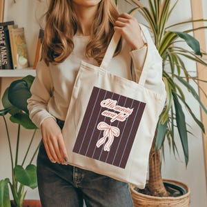 May include: A cream-coloured tote bag with a rectangular design. The design features the text "His mercies never fail" and a bow illustration, set against a background of brown and blue vertical stripes.