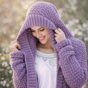 May include: A lavender hooded sweater with a textured knit design. The sweater features a large hood and long sleeves. The person in the image is wearing a white shirt underneath the sweater.