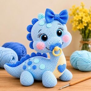 May include: A light blue crocheted dinosaur plush toy featuring a large blue bow on its head, a yellow pacifier in its mouth, pink cheeks, and a blue heart patch on its chest. The dinosaur has dark blue spikes along its back and light blue spots.