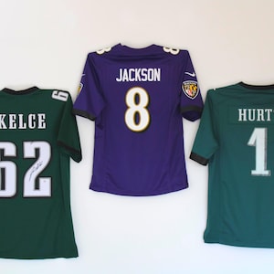 Peut inclure: Trois maillots de football accrochés à un mur blanc. Le maillot de gauche est vert avec des lettres blanches et le numéro 62. Le maillot du milieu est violet avec des lettres blanches et le numéro 8. Le maillot de droite est vert avec des lettres blanches et le numéro 1.