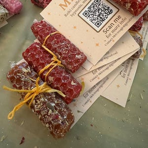 May include: Close-up of several handmade candles in shades of red and brown, tied with yellow twine. The candles are rectangular and textured, some with embedded herbs. Each candle is accompanied by a small card with the text "MELVI" and a QR code.