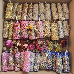 Puede incluir: Una colección de velas de pilar en una caja de cartón. Las velas cilíndricas están decoradas con flores y hierbas secas en varios colores, incluyendo rosa, amarillo y azul. Las velas están dispuestas en filas ordenadas, creando una exhibición visualmente atractiva.