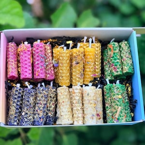 Puede incluir: Una caja de velas cilíndricas coloridas. Las velas son en tonos de rosa, dorado, verde, azul y crema, y están decoradas con flores secas y purpurina. La caja tiene un interior de colores del arcoíris y está llena de papel arrugado negro.