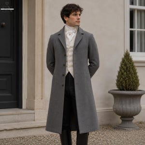 Könnte beinhalten: Ein langer, grauer Mantel mit maßgeschneiderter Passform, getragen über einer cremefarbenen Weste und einem weißen Jabot. Der Mantel hat ein klassisches Design mit Knopfverschluss. Das Outfit wird durch eine schwarze Hose und Stiefel ergänzt, was einen formellen, historischen Look erzeugt.