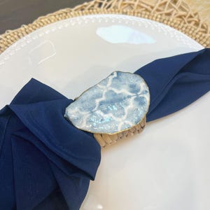 May include: A navy blue napkin is held by a decorative napkin ring. The ring features a light blue and white design with gold trim, set on a woven base. The napkin and ring are placed on a white plate.