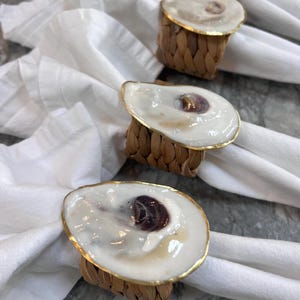 May include: Set of four oyster shell napkin rings with a gold trim and woven rattan base. The oyster shells are white with a pearlescent finish and a dark purple interior.