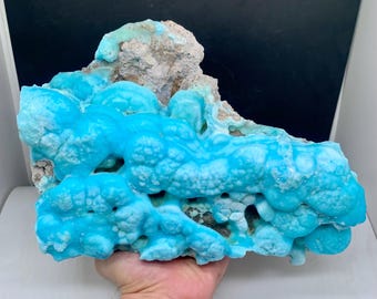May include: A large, irregularly shaped mineral specimen featuring a vibrant, turquoise-blue color. The surface has a bubbly, textured appearance with areas of light brown and beige. The mineral is held in a hand, showcasing its size and unique form.