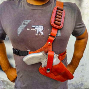 Può includere: Una fondina da spalla in pelle marrone con un porta cartucce abbinato. La fondina è progettata per trasportare una pistola d'argento. La pelle è di un ricco colore marrone-rossastro, con cuciture visibili e hardware in metallo.