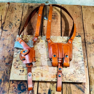 Puede incluir: Un portafusil de cuero marrón con una funda para cargador a juego. El portafusil tiene un diseño moldeado para adaptarse a un arma de fuego, con correas ajustables y herrajes metálicos. El cuero es de color marrón cálido.