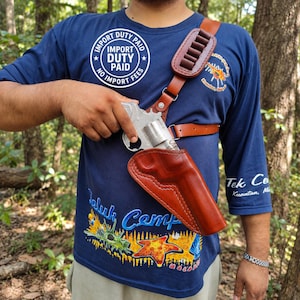 May include: A brown leather shoulder holster with a matching ammunition carrier strap featuring six loops. The holster is worn over a navy blue t-shirt with graphic prints and text, including a circular logo that reads IMPORT DUTY PAID.