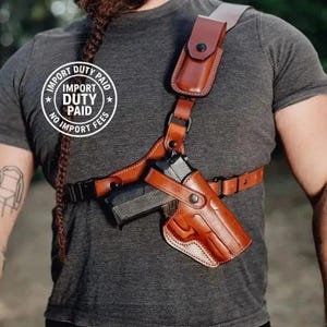 Peut inclure: Un système d'étui d'épaule en cuir marron comprenant une pochette moulée pour arme de poing, un porte-chargeur intégré avec fermeture à pression, des sangles réglables et des pièces métalliques, porté sur un t-shirt gris foncé.
