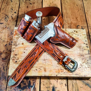 Puede incluir: Un conjunto de cinturón y funda de cuero marrón con intrincados diseños florales. El conjunto incluye un cinturón con hebilla plateada, una funda y dos bolsas para cargadores. El arma es plateada. El conjunto se exhibe sobre una superficie de madera.