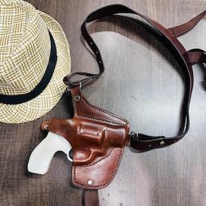 Może przedstawiać: Brązowa skórzana kabura z białym pistoletem w środku. Kabura jest przymocowana do brązowego skórzanego paska na ramię.