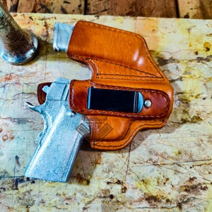 Op de afbeelding: Een bruin leren holster met een zwarte clip, met een zilveren pistool. Het holster is gestikt met donker draad en heeft een gebogen ontwerp. Het pistool is gedeeltelijk zichtbaar, met een zilveren afwerking.