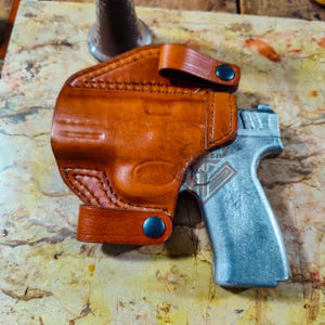 Puede incluir: Una funda de cuero marrón con una pistola plateada. La funda está cosida y tiene un cierre de botón a presión. La pistola está parcialmente insertada, mostrando su acabado metálico.