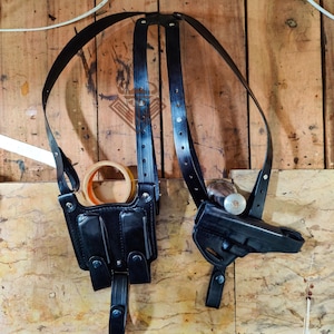 May include: Black leather shoulder holster with two pouches and straps. The holster is designed to hold a handgun and is suspended by adjustable straps. The background is a wooden surface.