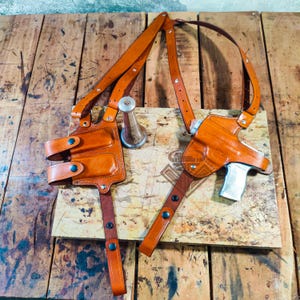 Puede incluir: Una funda de hombro de cuero marrón con dos bolsas y una funda para pistola. La funda tiene correas ajustables y herrajes metálicos. El cuero es de un color marrón rojizo cálido, y el diseño es visible sobre un fondo de madera.