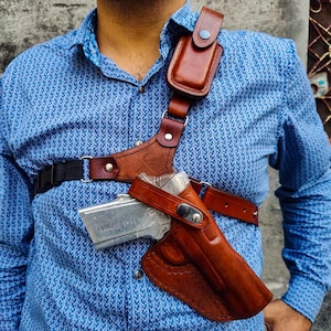 Op de afbeelding: Een bruin leren schouderholster met een bijpassend leren etui en een zwarte nylon band. Het holster is ontworpen om een ​​handvuurwapen te dragen. De persoon draagt ​​een blauw overhemd met patroon.