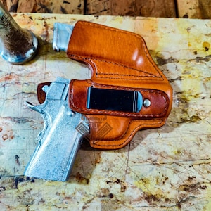 Puede incluir: Una funda de cuero marrón con un clip negro, que sostiene una pistola plateada. La funda está cosida con hilo oscuro y tiene un diseño curvo. La pistola es parcialmente visible, con un acabado plateado.