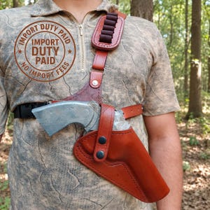 May include: A brown leather shoulder holster with a silver-colored revolver, featuring a shoulder strap with ammunition loops, a chest strap, and a snap-button closure. The image includes the text IMPORT DUTY PAID NO IMPORT FEES.