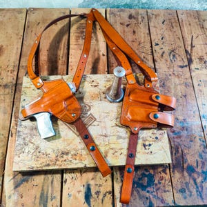 以下が含まれることがあります： 銀色の拳銃が付いた茶色の革製ショルダーホルスター。ホルスターは調節可能なストラップとダブルマガジンポーチを備えています。革は暖かく、ミディアムブラウン色で、金具はシルバーです。