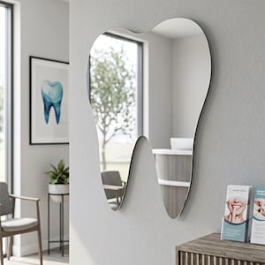 May include: A unique, tooth-shaped mirror hangs on a gray wall. The mirror reflects a room with a window, chair, and a framed dental illustration. A small plant and brochures are visible nearby, suggesting a dental office setting.