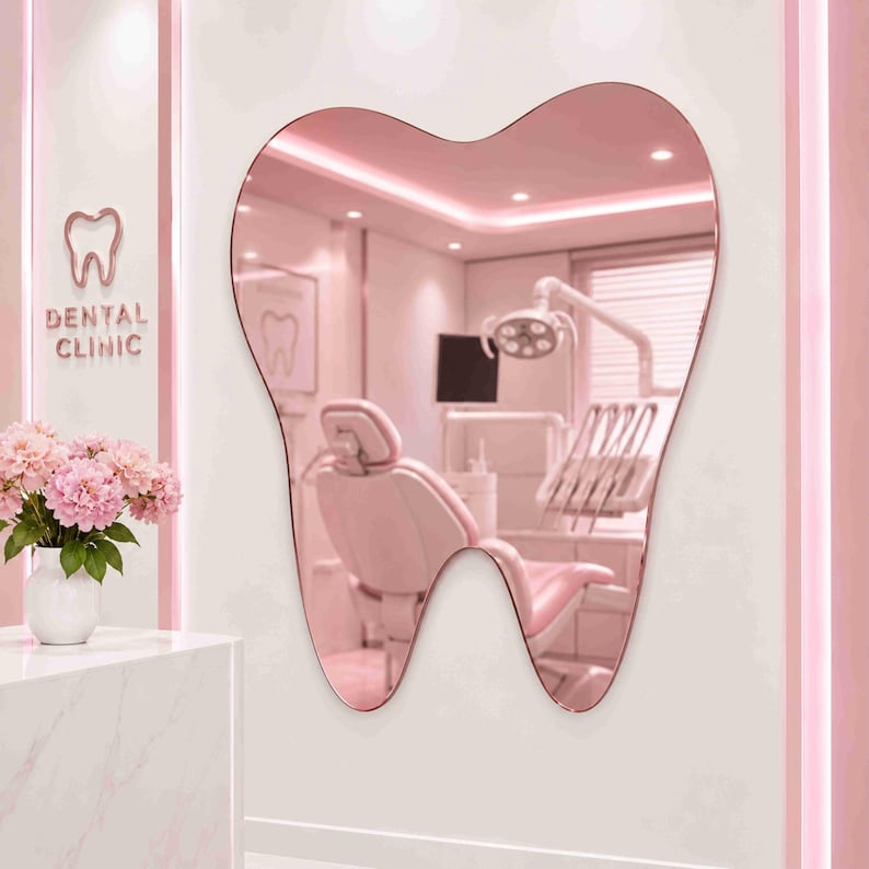 May include: A wall-mounted mirror shaped like a tooth with a rose-gold tinted reflective surface. The mirror reflects a dental office interior featuring a pink dental chair, medical lighting, and equipment. A white vase with pink flowers sits nearby.