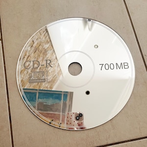 May include: A reflective CD-R disc with the text "CD-R Compact Disc Rewritable High Speed" and "700 MB" printed on the surface. The disc reflects the surrounding environment, including a building and a phone.