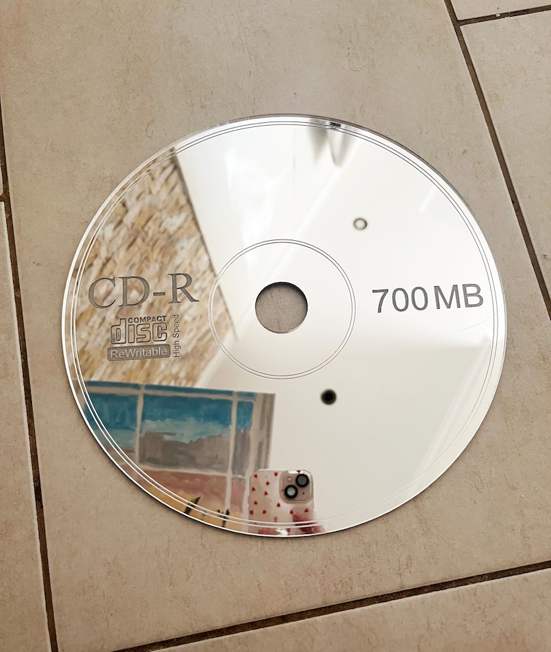 May include: A reflective CD-R disc with the text "CD-R Compact Disc Rewritable High Speed" and "700 MB" printed on the surface. The disc reflects the surrounding environment, including a building and a phone.