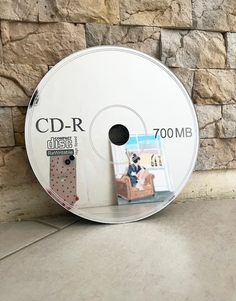 May include: A reflective CD-R disc with the text "CD-R" and "700 MB" printed on the surface. The disc reflects an image of a person sitting in a chair. The disc is sitting on a tiled surface.