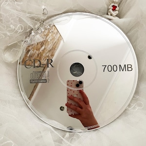 May include: A reflective CD-R disc with the text "CD-R COMPACT disc ReWritable" and "700 MB". The disc reflects a hand holding a phone with a heart pattern. A small figurine of an angel holding a heart is in the background.