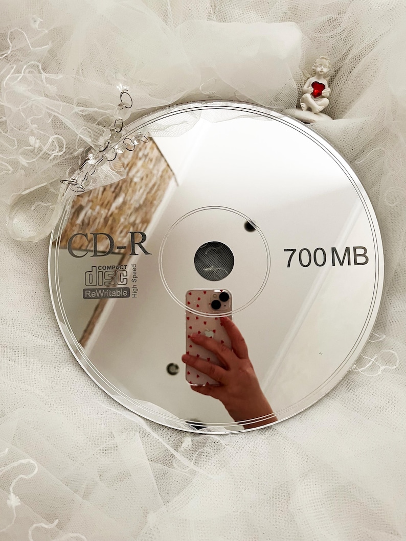 May include: A reflective CD-R disc with the text "CD-R COMPACT disc ReWritable" and "700 MB". The disc reflects a hand holding a phone with a heart pattern. A small figurine of an angel holding a heart is in the background.