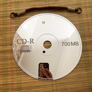 May include: A reflective CD-R disc with the text "CD-R" and "700 MB" printed on it. A brown and tan hair tie is above the disc. The disc reflects the image of a person holding a phone.