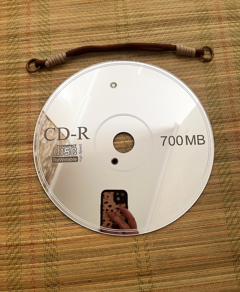 May include: A reflective CD-R disc with the text "CD-R" and "700 MB" printed on it. A brown and tan hair tie is above the disc. The disc reflects the image of a person holding a phone.