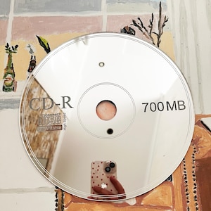 May include: A reflective silver CD-R disc, displaying the text "CD-R" and "700 MB". The disc reflects the surrounding environment, including a person holding a phone with a heart pattern.