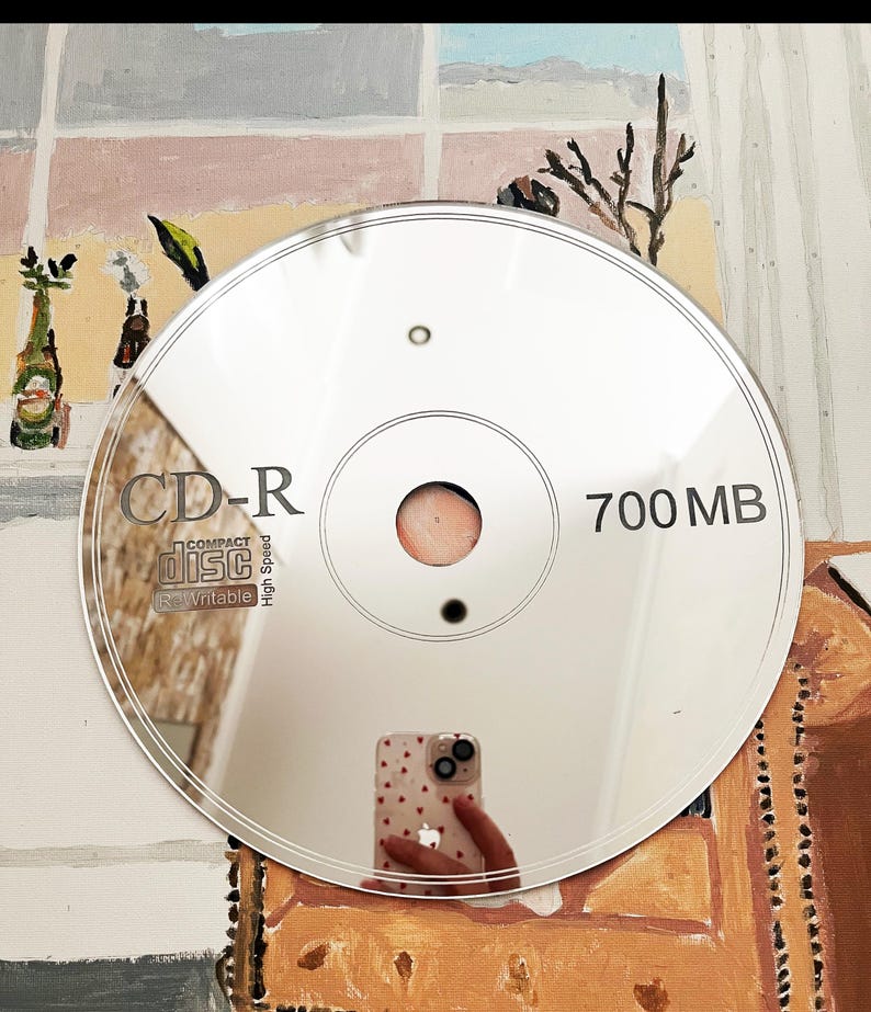 May include: A reflective silver CD-R disc, displaying the text "CD-R" and "700 MB". The disc reflects the surrounding environment, including a person holding a phone with a heart pattern.