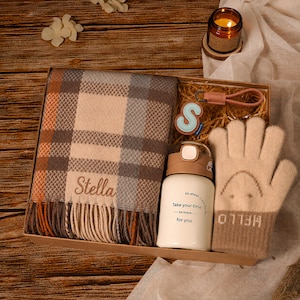 Könnte beinhalten: Eine Geschenkbox mit einem karierten Schal, auf dem der Name Stella eingestickt ist, einem beigen Paar Strickhandschuhe mit einem Smiley und dem Wort HELLO, einer weißen Isolierflasche mit dem Text Take your time be brave for you und einem S-Schlüsselanhänger.
