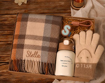 May include: A gift box containing a plaid scarf with the name Stella embroidered on it, a beige pair of knit gloves with a smiley face and the word HELLO, a white insulated bottle with the text Take your time be brave for you, and a letter S keychain.