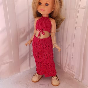 May include: A doll wearing a handmade, bright red crochet halter top and matching skirt. The skirt has an openwork design. The doll has blonde hair, blue eyes, and is wearing a floral headband and beige shoes.